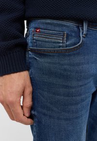 Mėlynos džinsinės kelnės su nublukusiu finišu, puošniu oranžiniu siuvinėjimu ir maža raudona etikete šalia monetų kišenės. Rankos pozicija ant kojos.