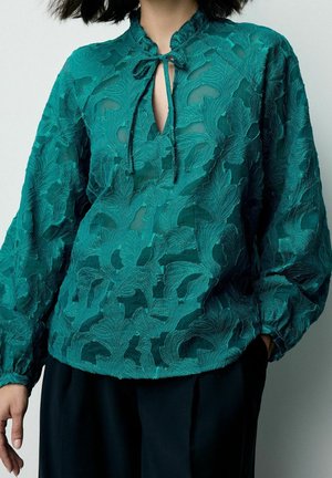 Femme portant un chemisier en dentelle florale transparente bleu sarcelle avec col goutte d'eau et lien, associé à un pantalon noir, sur fond uni.