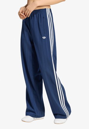 Persona che indossa pantaloni della tuta Adidas blu navy con strisce bianche e logo, abbinati a scarpe bianche, in piedi davanti a uno sfondo semplice.