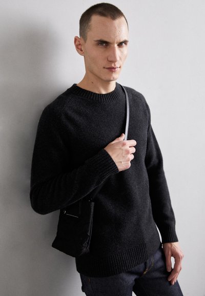 Mørkegrå strikkegenser med ribbestrikket halslinning og mansjetter, kombinert med en liten svart crossover-bag. Modellen står mot en lys vegg.