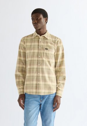 Jeune homme portant une chemise à carreaux beige et vert clair à boutons avec manches longues et un jean bleu, debout devant un fond blanc uni.