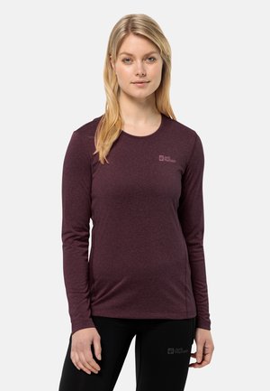 Blonde vrouw in een strak, lange mouw, donkerpaarse top en zwarte broek, staand tegen een effen lichte achtergrond.