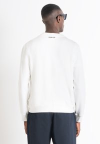 Sudadera blanca con puños y dobladillo acanalados. Presenta un cuello redondo y un logo en la parte posterior. Textura suave y corte relajado. Lleva puesta con pantalones azul marino.