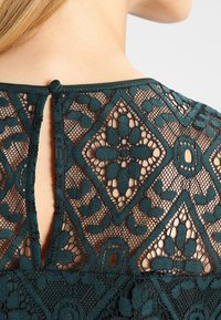 Tela de encaje en color teal con intrincados patrones de diamantes y flores; cuenta con un escote en forma de llave en la parte posterior con un pequeño cierre de botón en el cuello.