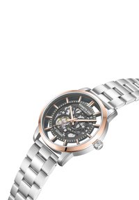 Automatisk klocka med transparent svart urtavla, silver- och roséguldfärger, samt ett silverfärgat metallarmband. Har synliga kugghjul och intrikata detaljer.