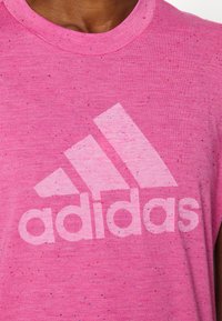 Camiseta deportiva rosa con una superficie texturizada, que presenta un logo "adidas" en rosa claro y motas de azul y rojo distribuidas por toda la tela.