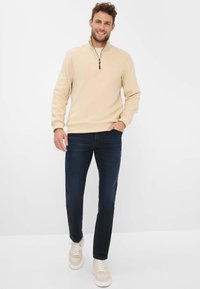 Beige ritssluiting fleece sweatshirt met een staande kraag, gecombineerd met donkerblauwe jeans en lichtgekleurde sneakers, staand tegen een effen achtergrond.