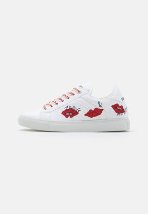 Zapatilla blanca de caña baja con labios rojos de purpurina, texto manuscrito en negro y cordones con patrón rojo y blanco sobre un fondo blanco.