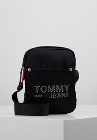 Borsa a tracolla nera in tessuto con parte anteriore in rete, caratterizzata dal logo "TOMMY JEANS" in testo riflettente argento. Include una tracolla regolabile e un accento rosso.