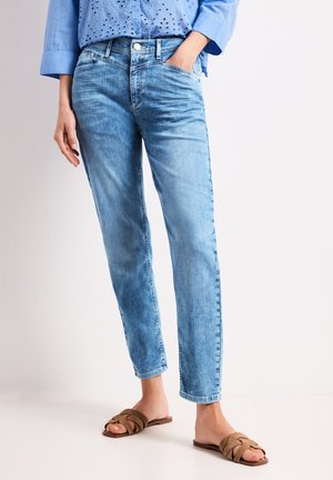 Kobieta w jasnoniebieskich jeansach o prostych nogawkach, jasnoniebieskiej bluzce z oczkami i brązowych płaskich sandałach, stojąca z jedną ręką w kieszeni.