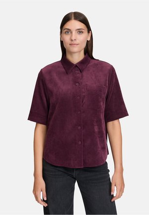 Donna che indossa una camicia in velluto a coste bordeaux con maniche corte e bottoni, dotata di una tasca sul petto, e jeans scuri, in piedi contro uno sfondo neutro.