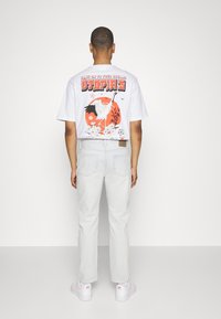 Mężczyzna odwrócony tyłem, ubrany w białą koszulkę z grafiką przedstawiającą żurawia oraz tekst, jasnoszare dżinsy i białe trampki z różowymi akcentami.