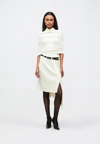 Λευκό υφασμάτινο μπλουζάκι με ψηλό γιακά, συνδυασμένο με κρέμ φούστα τύπου περιτυλίγματος με μπροστινή σχισμή και μαύρη ζώνη, φορεμένο με μαύρες καλσόν και τακούνια.