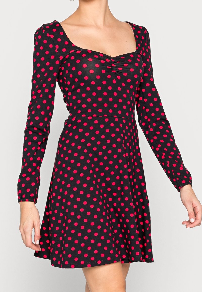 Frau in einem schwarzen langärmeligen Kleid mit roten Polka-Dots und einem gerafften Herzausschnitt, die mit leicht erhobenen Armen steht.