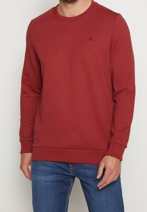 Mand iført en langærmet, rundhalset vinrød sweatshirt med en lille broderet logo på brystet, kombineret med blå jeans.