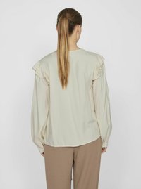 Lys beige langærmet bluse med volants detaljer på skuldrene, blød tekstur, afrundet kant, båret med lysebrune bukser, set fra ryggen.