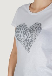 Valge T-särk, millel on sädelev südame muster, mis koosneb mustadest ja hõbedast nupudest, keskel "Liu Jo" tekst. Lühikesed varrukad, ümmargune kaelus.