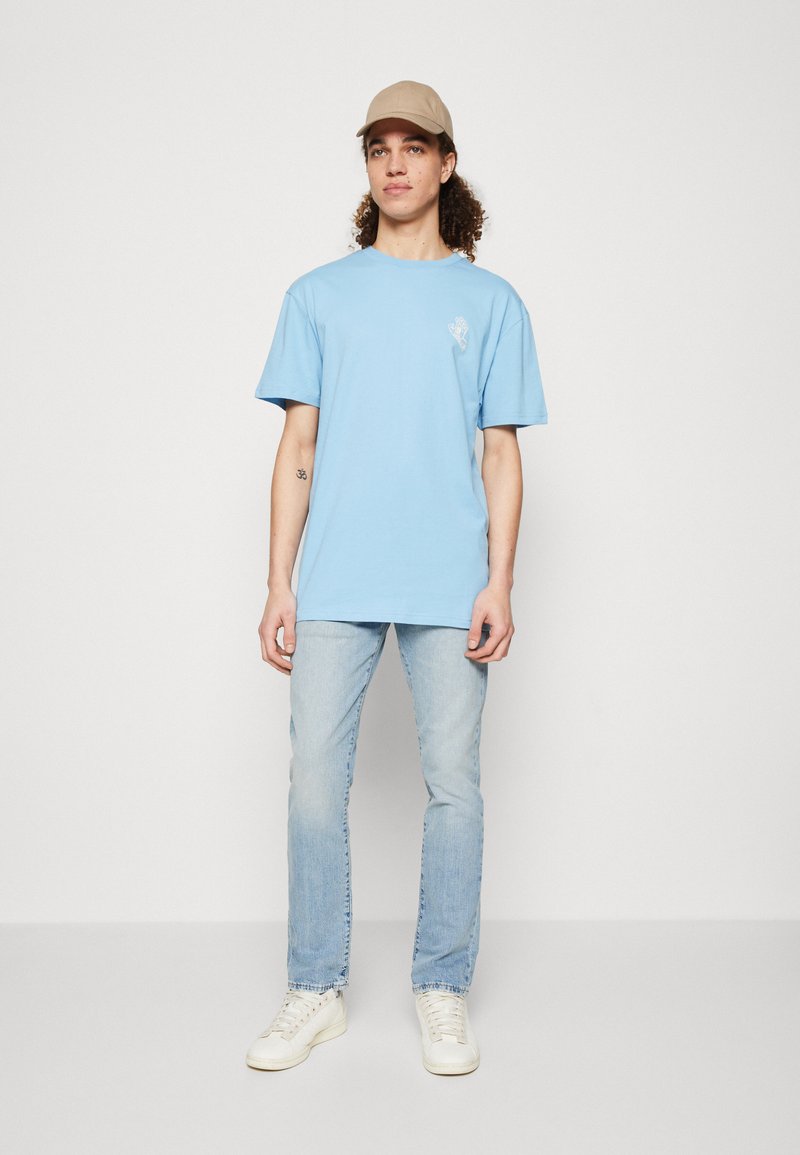 Giovane con capelli ricci che indossa un cappellino beige, una maglietta grafica azzurro chiaro, jeans chiari e sneaker bianche, in piedi davanti a uno sfondo semplice.