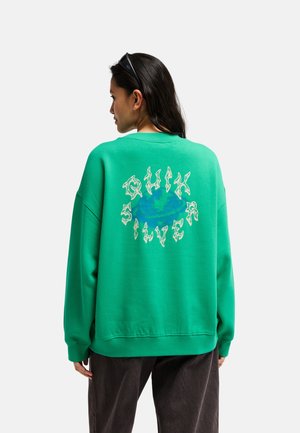 Vrouw draagt groen sweatshirt met gestileerde "Quiksilver" tekst en wereldbolgrafiek op de rug, gecombineerd met donkere broek en zonnebril op haar hoofd.