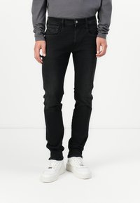 Black slim-fit jeans with a slightly faded finish, five pockets, and a metal button closure, worn with white sneakers and gray long-sleeve shirt.