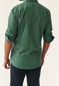 Camisa de cuadros verdes de botones con mangas remangadas, que presenta una textura suave y un corte estructurado. La vista trasera muestra el diseño de la yugo y el dobladillo.