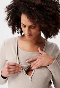 Vrouw met krullend haar die een beige knoopjes-top opent, waarbij ze een binnenlaag onthult, terwijl ze naar haar handen kijkt.