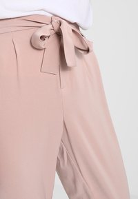 Vaaleanpunaiset, korkeavyötäröiset housut, joissa on solmioneula vyötäröllä. Pehmeä, sileä kangas, jossa hienovaraiset laskokset ja sivutaskut.