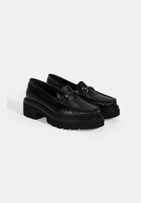 Zwarte leren loafers met een ronde neus, textuurdetails, metalen kettingaccenten en een stevige zwarte zool. Plat ontwerp voor gemakkelijk dragen.