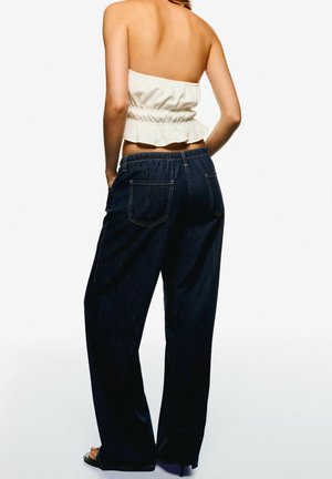 Kobieta ubrana w biały, bezramkowy top i luźne ciemnoniebieskie dżinsy, stojąca z rękami w kieszeniach, tyłem do widza.