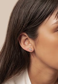 Silver stud earrings featuring a skull and crossbones design, with a polished finish and small gemstones. Worn on a model's ear.