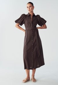 Hnědé šaty na knoflíky s páskem v pase, krátkými puff rukávy a k délce po kolena. Materiál vypadá texturovaně a lehce.
