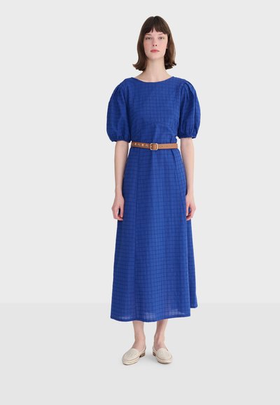 Femme portant une robe midi à carreaux bleus avec des manches bouffantes, une ceinture marron et des chaussures crème sans lacets, debout devant un fond uni.