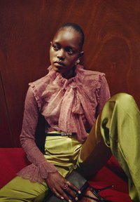 Blusa de encaje en suave rosa con cuello y mangas con volantes, combinada con pantalones de satén verde oliva. Un bolso de mano negro y anillos adornan las manos.