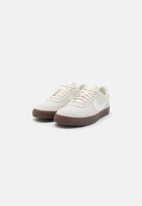 Graue Wildledersneaker mit einem weißen Swoosh-Logo, flachen Schnürsenkeln, gepolstertem Zungenbereich und einer strukturierten, braunen Gummisohle.