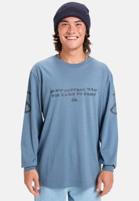 Camisa azul de manga larga con un eslogan gráfico en gris oscuro, que presenta un corte holgado y puños acanalados. Pantalones de mezclilla casuales y un gorro navy.