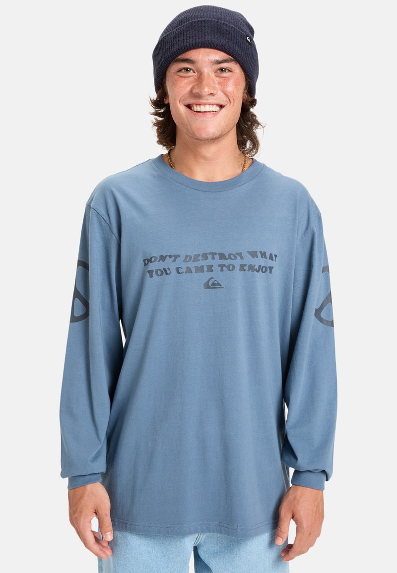 Camisa azul de manga larga con un eslogan gráfico en gris oscuro, que presenta un corte holgado y puños acanalados. Pantalones de mezclilla casuales y un gorro navy.