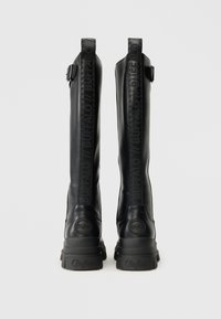 Fekete bőr, térd fölé érő csizmák, amelyek robusztus talppal, a hátsó részen dombornyomott márka logóval és az oldalon egy csattal rendelkeznek.