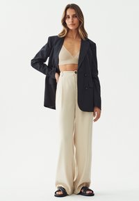 Crni oversized sako, bež rebrasti bralette, kremaste široke hlače i crne sandale na klizanje, prikazujući minimalistički, slojeviti izgled.