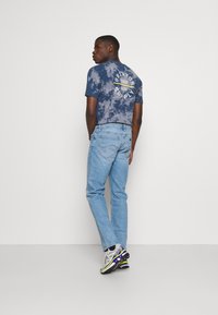 Camiseta de telaraña en color navy con branding blanco en la parte trasera, combinada con jeans de color azul claro y zapatillas plateadas con acentos en negro y púrpura.