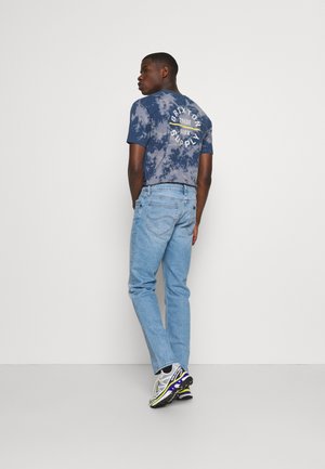 Marinblåa tie-dye T-shirt med vit branding på ryggen, kombinerad med ljusblå jeans och silverfärgade sneakers med svarta och lila detaljer.