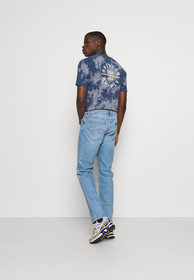 Camiseta de telaraña en color navy con branding blanco en la parte trasera, combinada con jeans de color azul claro y zapatillas plateadas con acentos en negro y púrpura.