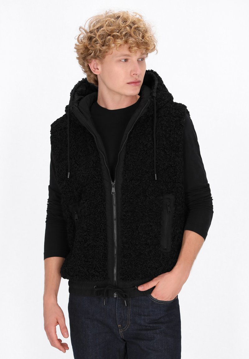 Černá bez rukávů s kapucí, s texturovanou látkou, zipem v přední části, nastavovací šňůrkou na dolním lemu a bočními kapsami. Nositá přes černé tričko s dlouhým rukávem.