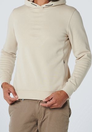 Man wearing beige hooded sweatshirt with subtle hexagonal embossed design on chest and light brown pants, adjusting hem.