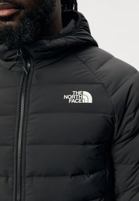 Giacca nera imbottita con zip frontale. Presenta un cappuccio e un logo bianco di "THE NORTH FACE" sul lato sinistro del petto. Texture liscia.