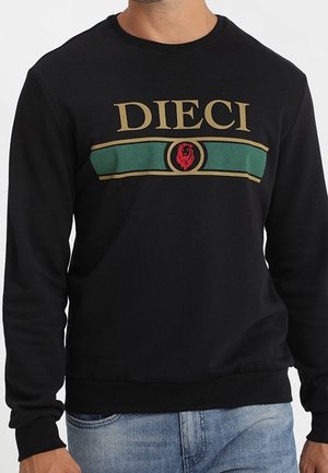 Zwarte sweater met lange mouwen met de tekst "DIECI" boven een groene streep en rood leeuws-embleem, gedragen door een persoon met een blauwe jeans.