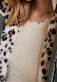 Beżowy prążkowany top na ramiączkach z falbankowym dekoltem, noszony pod zielonym płaszczem z kołnierzem z futra ekologicznego w panterkę.