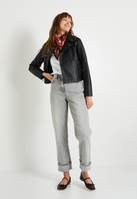 Femme portant une veste en cuir noir, une chemise blanche, une écharpe à motifs rouges, un jean gris clair retroussé et des ballerines noires, se tenant contre un mur blanc.