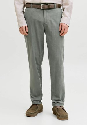 Mann iført lysegrå bukser, en beige skjorte med skjorten stoppet inn, et brunt flettet belte og brune semskede sko, står på hvit bakgrunn.