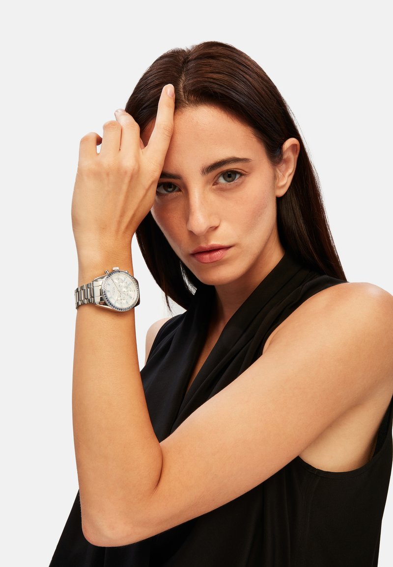 Montre chronographe argentée avec un bracelet en acier inoxydable, cadran blanc avec des accents et des index bleus, présentée sur un poignet contre un haut noir.