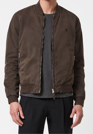 Mand iført en brun bomberjakke med lynlås over en mørkegrå T-shirt og sorte bukser mod en ensfarvet baggrund.